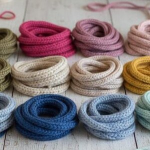 May include: A collection of twelve coiled bundles of braided cord in various colors, including olive green, magenta, mauve, pale pink, beige, cream, mustard yellow, light blue, navy blue, and mint green, arranged in a grid pattern.