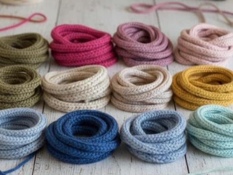 May include: A collection of twelve coiled bundles of braided cord in various colors, including olive green, magenta, mauve, pale pink, beige, cream, mustard yellow, light blue, navy blue, and mint green, arranged in a grid pattern.