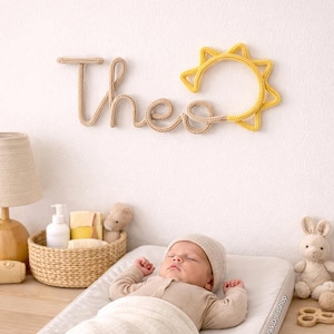 May include: A wall-mounted name sign made of beige knitted yarn forming the letters 'Theo' with a yellow knitted sun icon attached to the end. The letters are connected in a continuous cursive script style, creating a decorative wall hanging.