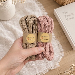 May include: Two bundles of thick, braided yarn cords in beige and dusty pink. Each bundle is secured with a tan paper label featuring the text "handmade with love" and a small red heart icon. The cords have a textured, knit-like appearance.