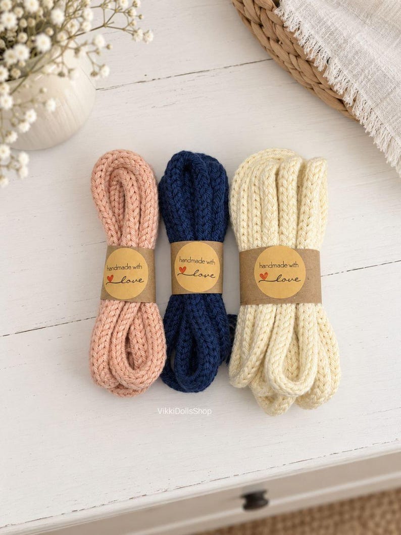 May include: Three bundles of thick, knitted cord in soft pink, navy blue, and cream. Each bundle is secured with a brown paper band featuring the printed text 'handmade with love' and a small red heart icon. The cords have a textured, braided appearance.