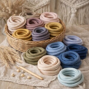 May include: A collection of coiled cotton cord in various colors including beige, pink, yellow, purple, olive green, and shades of blue, arranged in a woven basket and on a fabric surface with a wooden crochet hook and small wooden beads.