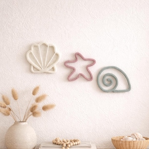 May include: Three wall-mounted decorative shapes made of yarn, including a cream-colored scallop shell, a dusty pink starfish, and a sage green spiral seashell. These items are arranged horizontally on a textured wall above a vase and a basket.