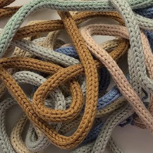 May include: A collection of intertwined knitted cords in various muted colors including mustard yellow, sage green, soft blue, and pale pink. The cords feature a distinct braided texture and are arranged in a tangled, overlapping pattern.