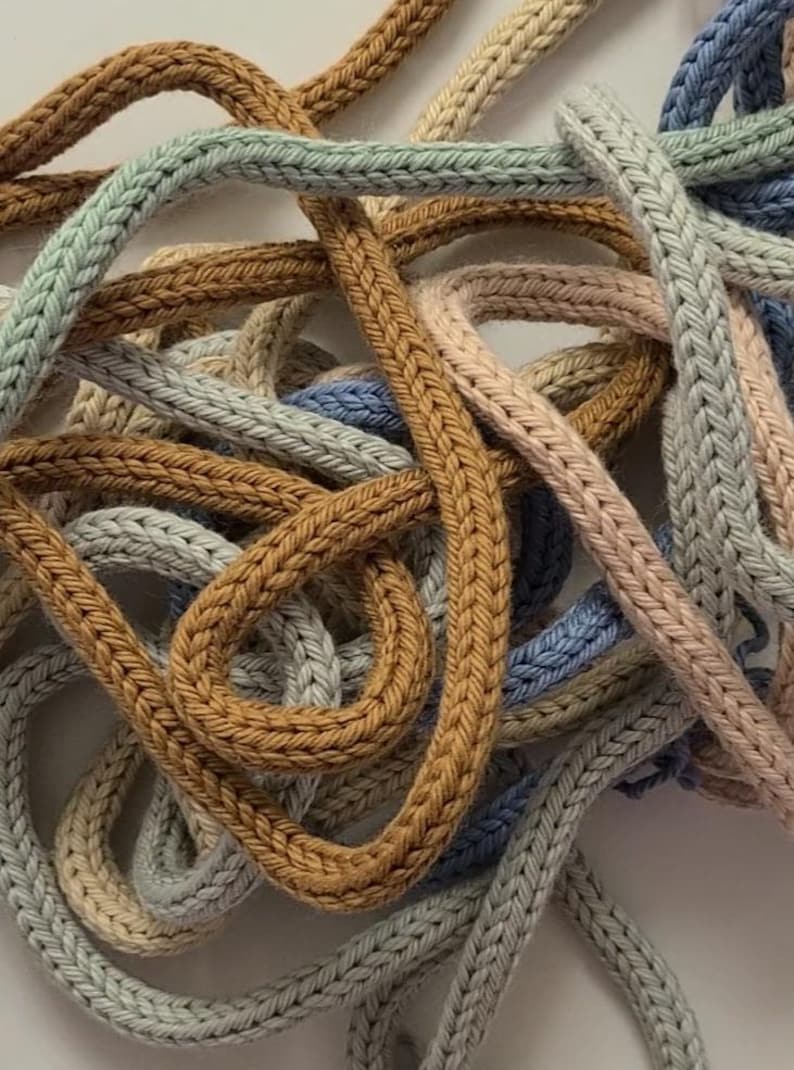 May include: A collection of intertwined knitted cords in various muted colors including mustard yellow, sage green, soft blue, and pale pink. The cords feature a distinct braided texture and are arranged in a tangled, overlapping pattern.