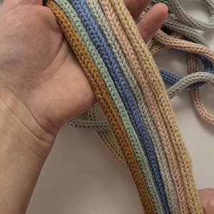 May include: A collection of multi-colored knitted cords featuring a braided texture. The cords are presented in a palette of soft pastel and earthy tones, including shades of mustard yellow, sage green, light blue, beige, and dusty pink.