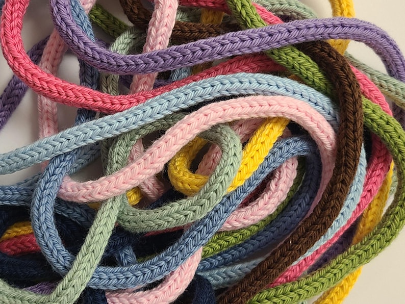 May include: A collection of multi-colored knitted cords tangled together. The cords feature a braided texture and come in various shades including pink, light blue, lavender, yellow, sage green, dark brown, and navy blue.