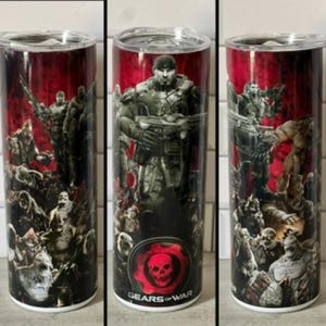 May include: A stainless steel tumbler featuring a Gears of War design. The design includes characters, a skull emblem, and the text "Gears of War" against a red and black background. The tumbler has a clear lid.