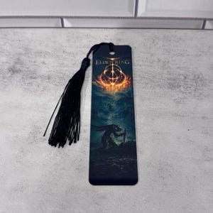 May include: A rectangular bookmark featuring the Elden Ring logo at the top, with a dark, atmospheric illustration of a figure kneeling in a landscape beneath a glowing golden symbol. A black tassel is attached to the top through a small hole.