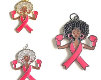 May include: A pink enamel charm with a black woman wearing boxing gloves and a pink ribbon. The charm is made of metal and has a sparkly finish.