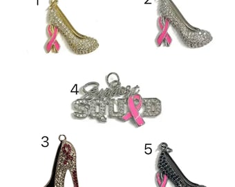 May include: Five charm pendants featuring a pink ribbon and a high heel shoe design. The charms are made of silver and gold metal and are decorated with rhinestones. One charm features the words "Support Squad" with a pink ribbon.