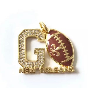 Pu&ograve; includere: Ciondolo in oro con una grande lettera G ricoperta di strass e un pallone da football con un design a giglio. Il ciondolo dice "NEW ORLEANS" sotto la lettera G.