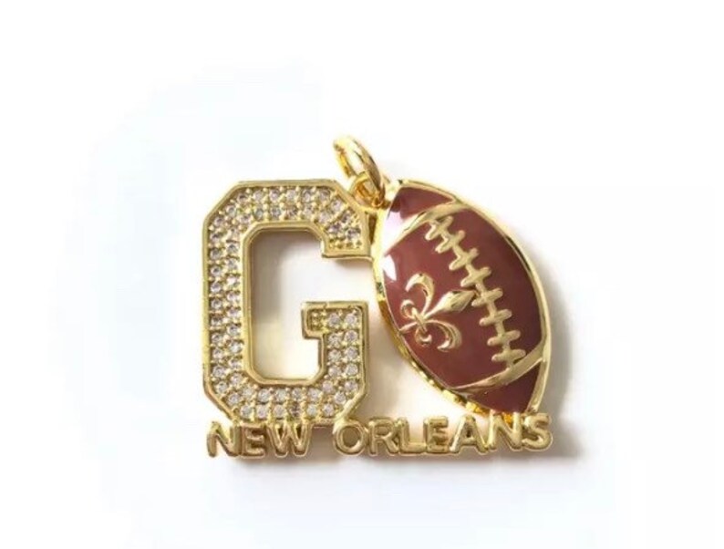 Pu&ograve; includere: Ciondolo in oro con una grande lettera G ricoperta di strass e un pallone da football con un design a giglio. Il ciondolo dice "NEW ORLEANS" sotto la lettera G.