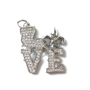 Pu&ograve; includere: Un ciondolo in argento con la parola "LOVE" scritta in grandi lettere, ogni lettera ricoperta di piccoli strass trasparenti. Una fleur-de-lis argentata si trova tra la "O" e la "V".