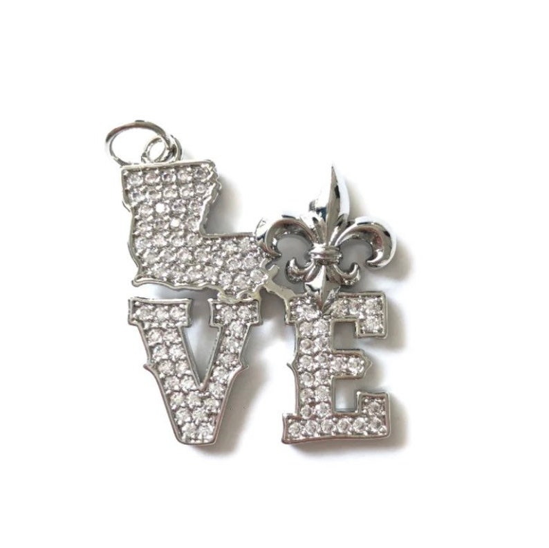 Pu&ograve; includere: Un ciondolo in argento con la parola "LOVE" scritta in grandi lettere, ogni lettera ricoperta di piccoli strass trasparenti. Una fleur-de-lis argentata si trova tra la "O" e la "V".