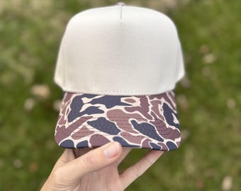 May include: A beige trucker hat with a camouflage pattern on the brim. The hat is held up against a blurred green background. The hat's crown is a solid beige color, and the brim features a pattern of brown, navy, and cream shapes.