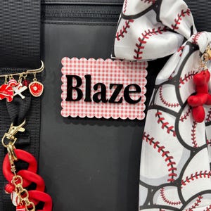 May include: A black bag featuring a scalloped rectangular name tag with a red and white gingham pattern and the name Blaze in black text. The bag is decorated with a baseball-themed fabric bow, a gold pin with baseball charms, and a red chain.