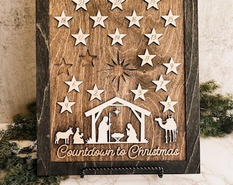 May include: Wooden Christmas advent calendar with a dark brown frame and a light brown interior. The calendar features star-shaped cutouts numbered 1-25, a nativity scene, and the text "Countdown to Christmas."