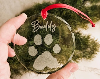 May include: Clear glass ornament with a paw print design and the name "Buddy" etched in script. The ornament has a red ribbon for hanging. The background includes green pine branches.