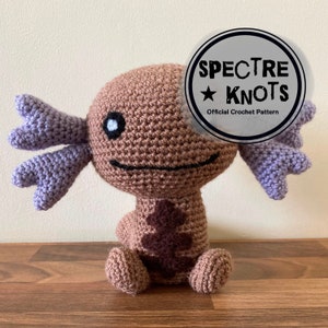 May include: A crocheted axolotl plush toy with brown and purple yarn. The toy has a brown body with a brown patch on its chest, purple gills, and black eyes. The toy is sitting on a wooden surface. The image includes the text "SPECTRE KNOTS Official Crochet Pattern".