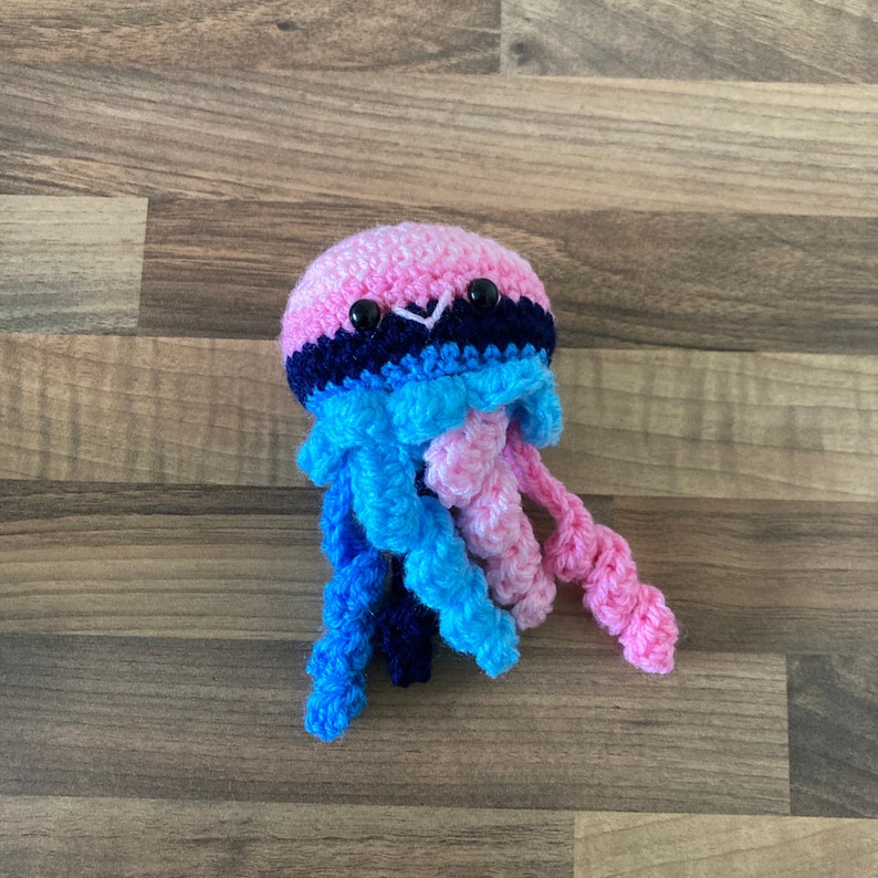 May include: A crocheted jellyfish toy with pink, blue and black stripes. The jellyfish has black eyes and a pink, blue and black striped body. The tentacles are pink, blue and black.
