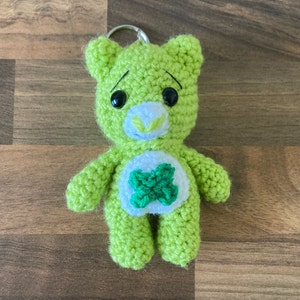 May include: Green crocheted Care Bear keychain with a green clover on its chest. The bear has black eyes and a white nose.