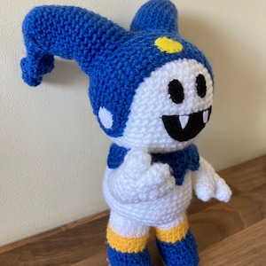 May include: A crocheted plush toy of a character with a blue and white hat, white body, and yellow accents. The character has a large smile with black teeth.