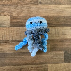 May include: A crocheted blue and grey jellyfish toy with white tentacles. The jellyfish has black eyes and a smiling mouth.