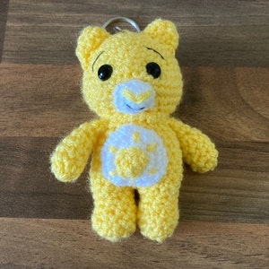 May include: Yellow crocheted Care Bear keychain with a white sun on its chest. The bear has black eyes and a black nose.