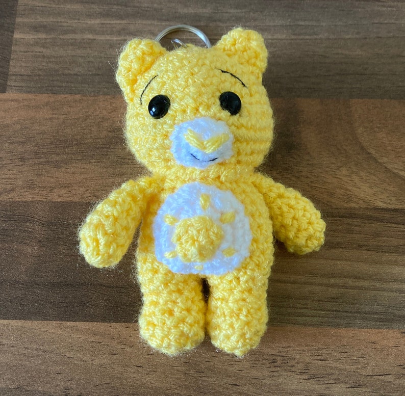 May include: Yellow crocheted Care Bear keychain with a white sun on its chest. The bear has black eyes and a black nose.