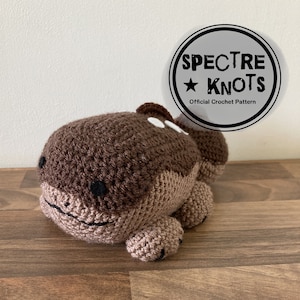 May include: A handmade crochet whale in shades of brown and tan, with black eyes and a smiling mouth. The whale has a rounded body and small fins. The image includes a circular logo that reads "SPECTRE KNOTS Official Crochet Pattern."