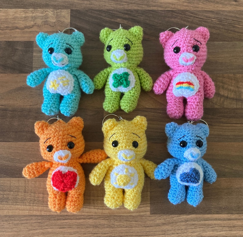 May include: Six crocheted Care Bear keychains in different colors: blue, green, pink, orange, yellow, and light blue. Each bear has a different symbol on its chest.