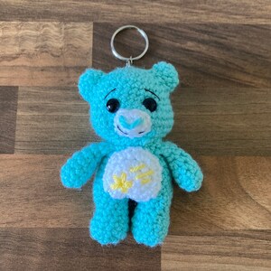 May include: A blue crocheted teddy bear keychain with a yellow flower on its chest. The bear has a white face with black eyes and a blue nose.