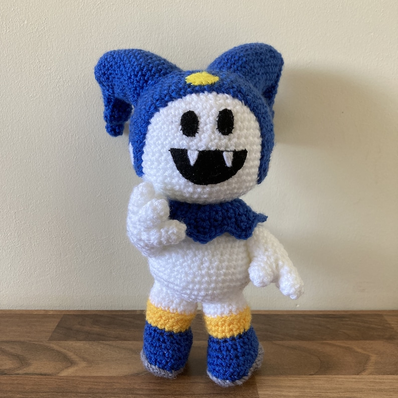 May include: A white crocheted plush toy with a blue hat and yellow and blue striped socks. The toy has a large, toothy grin and a yellow star on its hat.