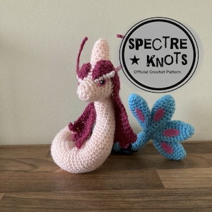 Puede incluir: Un caballito de mar de crochet rosa y azul con una melena rosa y una aleta de cola azul. El caballito de mar es un juguete de crochet hecho a mano con el texto "SPECTRE KNOTS Official Crochet Pattern" en un círculo negro.