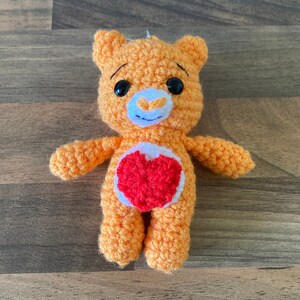 May include: A crocheted orange bear with a red heart on its chest. The bear has black eyes and a white nose.