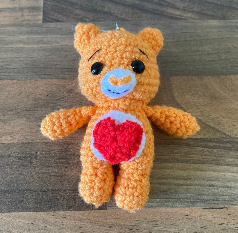 May include: A crocheted orange bear with a red heart on its chest. The bear has black eyes and a white nose.