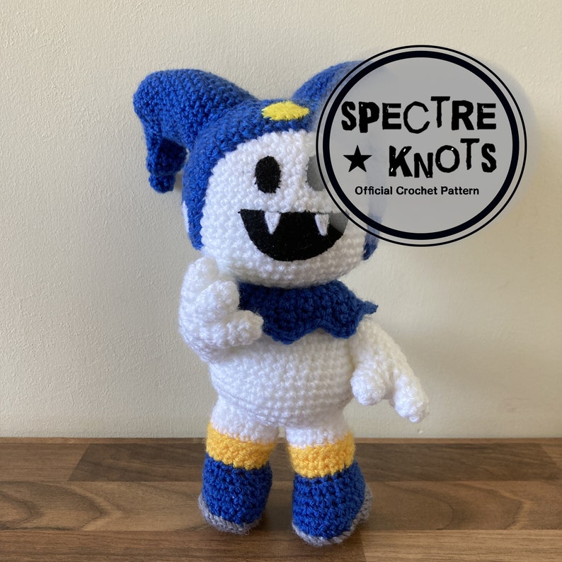 May include: A white crocheted plush toy with blue and yellow accents. The toy has a large smile with fangs and a blue jester hat. The toy is standing on a wooden surface. The text "SPECTRE KNOTS Official Crochet Pattern" is visible in a circle.