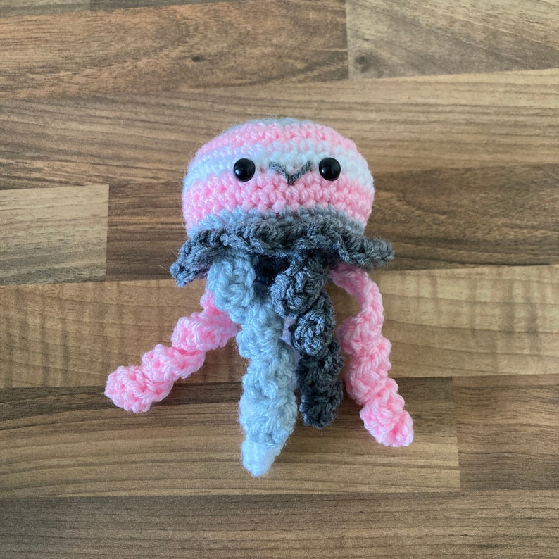 May include: A crocheted jellyfish toy with pink, white and grey stripes. The jellyfish has a small, round head with black eyes and a long, flowing body.