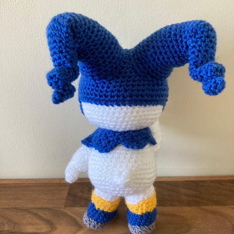 May include: A white crocheted stuffed toy with blue jester hat and yellow and blue striped legs. The toy is standing on a wooden surface.