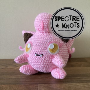 May include: A pink crochet plush toy with brown accents on the ears and a small mouth. The toy has large yellow and white eyes. The image includes a circular logo that says "Spectre Knots Official Crochet Pattern".