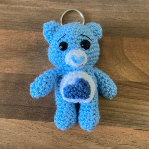 May include: A blue crocheted teddy bear keychain with a white heart on its chest. The heart has a blue cloud design. The bear has black eyes and a white nose.