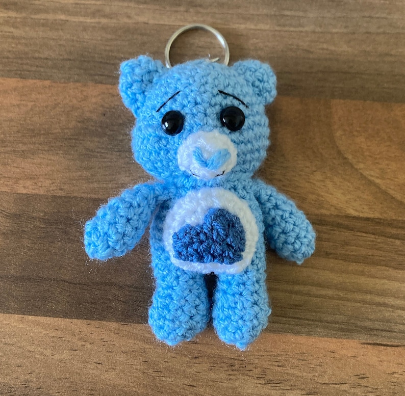 May include: A blue crocheted teddy bear keychain with a white heart on its chest. The heart has a blue cloud design. The bear has black eyes and a white nose.