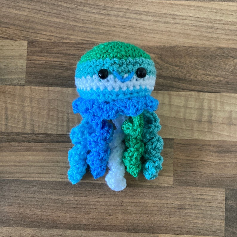 May include: A crocheted jellyfish toy in green, blue and white. The jellyfish has a smiling face with black eyes and a white mouth.