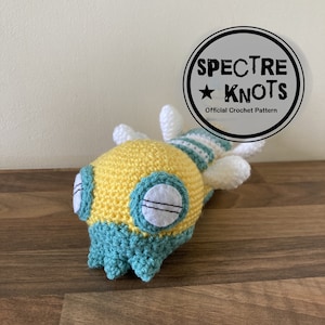 May include: A crocheted yellow and teal creature with white eyes and a striped tail. The creature is lying on a wooden surface. The text "SPECTRE KNOTS Official Crochet Pattern" is visible in a circle above the creature.