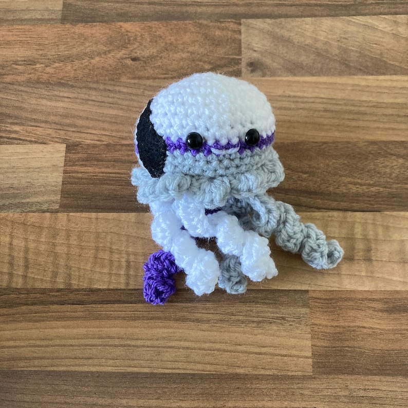 May include: A crocheted jellyfish toy with a white, grey, and purple colour scheme. The jellyfish has black eyes and a purple mouth.