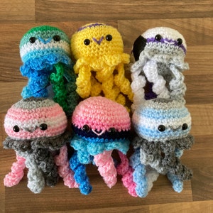May include: A set of ten crocheted jellyfish toys in various colours, including grey, pink, blue, yellow, and white. Each jellyfish has a different colour pattern and features a crocheted face with black eyes.