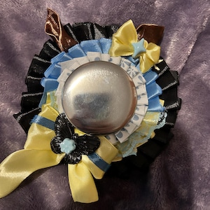 May include: A circular rosette badge featuring a central metallic silver button surrounded by layers of pleated black, light blue, and white polka-dot ribbon. The design includes yellow satin bows, a black butterfly charm, and light blue accents.