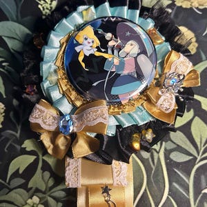 Puede incluir: Una insignia decorativa con una imagen circular central de personajes de anime. La insignia presenta capas de cintas negras, doradas y azul claro, encaje y lazos. Una gran gema azul está engastada en el centro de un lazo dorado.