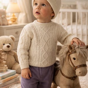 May include: A young child wearing a cream-colored cable-knit sweater and matching beanie, paired with lavender corduroy pants. The child is standing next to a rocking horse with a tan body and brown leather accents.